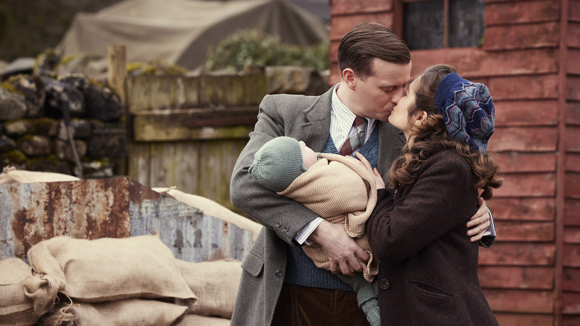 A man and woman share a kiss outside near a rustic wooden building. The man, dressed in a suit and tie, cradles a bundled baby in his arms. The woman, wearing a coat and a knitted hat, embraces him warmly.