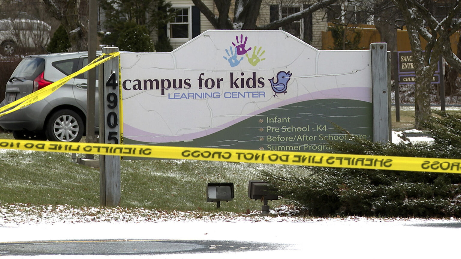 Plastic tape marked multiple times with the words Sheriff's Line Do Not Cross is attached to a wood post with the address number 4905 that is at one end of a sign with cracks in its paint that includes three handprints, an illustration of a bird and the words Campus for Kids, Infant, Pre-School - K4, Before/After School Care and Summer Programs that is placed in front of a pine landscaping shrub and stands on a small hill slope, with snow-dusted ground in the foreground and parked vehicles, tree trunks and a building in the background.