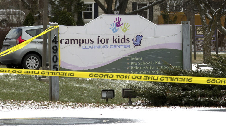 Plastic tape marked multiple times with the words Sheriff's Line Do Not Cross is attached to a wood post with the address number 4905 that is at one end of a sign with cracks in its paint that includes three handprints, an illustration of a bird and the words Campus for Kids, Infant, Pre-School - K4, Before/After School Care and Summer Programs that is placed in front of a pine landscaping shrub and stands on a small hill slope, with snow-dusted ground in the foreground and parked vehicles, tree trunks and a building in the background.