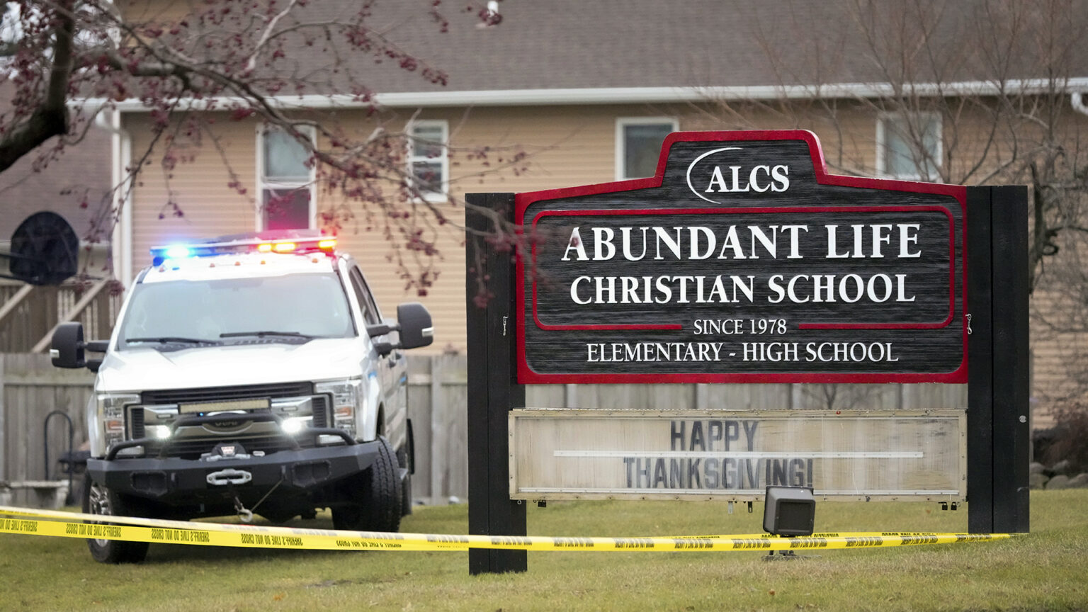 A police vehicle with illuminated siren lights is parked on a lawn next to a wood sign with the words ALCS, Abundant Life Christian School, Since 1978 and Elementary - High School, with a wood fence, multi-story house and trees in the background.