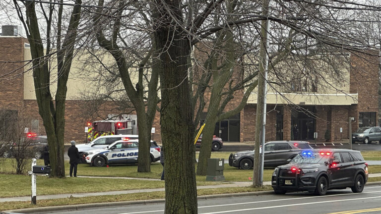 Multiple police vehicles with illuminated siren lights are parked on the side of a road and among leafless trees in a lawn in front of a multi-story brick building, with a leafless tree in the foreground.