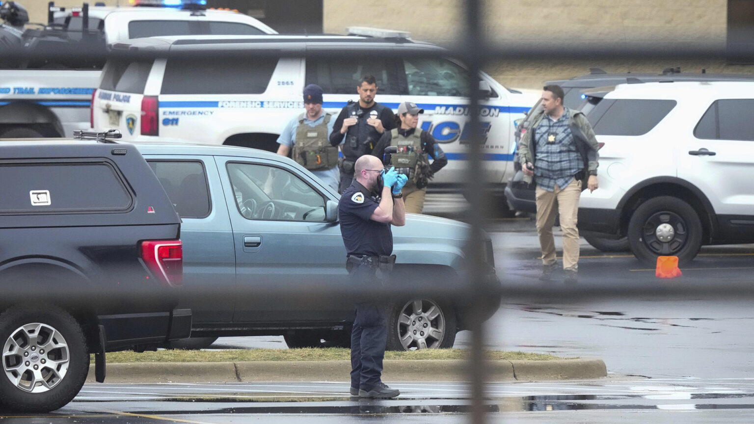 A Madison Police Department officer in uniform takes a photo while wearing gloves while standing in a parking lot among multiple parked vehicles, with other law enforcement officers wearing tactical vests and badges on lanyards walking in the background, with out-of-focus window blinds in the foreground.