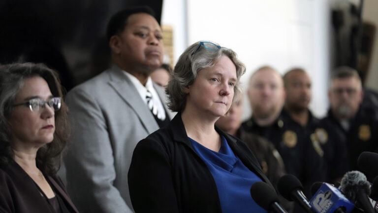 Satya Rhodes-Conway stands in front of multiple microphones with flags of different media organizations, with Melissa Agard standing to the side and Charles Tubbs and multiple uniformed law enforcement officers in the background.