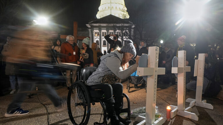 A person seated in a wheelchair writes on the surface of a piece of wood in the shape of a heart attached to the front of a wood cross standing on a wood mount next to another cross and a sign on a wood stand, with multiple people standing in the background, including a blurry person in motion, with two light sources on either side of a building with columns topped by a pediment and an illuminated dome.