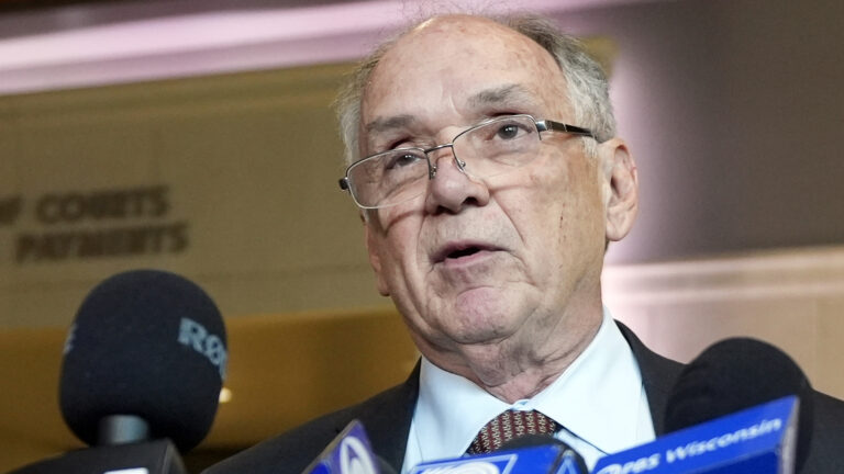 Jim Troupis speaks into a row of microphones with the flags of different media organizations inside a room with recessed lighting in the background.