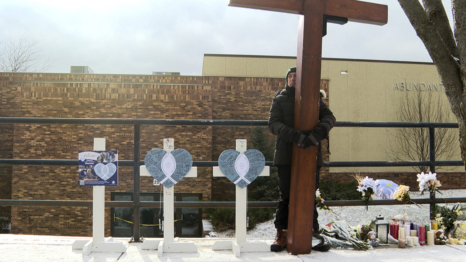 Dan Beazley holds a tall wood cross while standing in the middle of a makeshift memorial that includes two short wood crosses with front panels in the shape of a heart surrounding a vertical ichthys symbol and covered with handwritten notes next to a sign mounted on a wood stand on one side and  a series of flower bouquets and devotional candles on the other in front of a metal walkway fence, with a tree trunk, snow covered lawn, and multi-section building with a brick wall on one portion in the background.