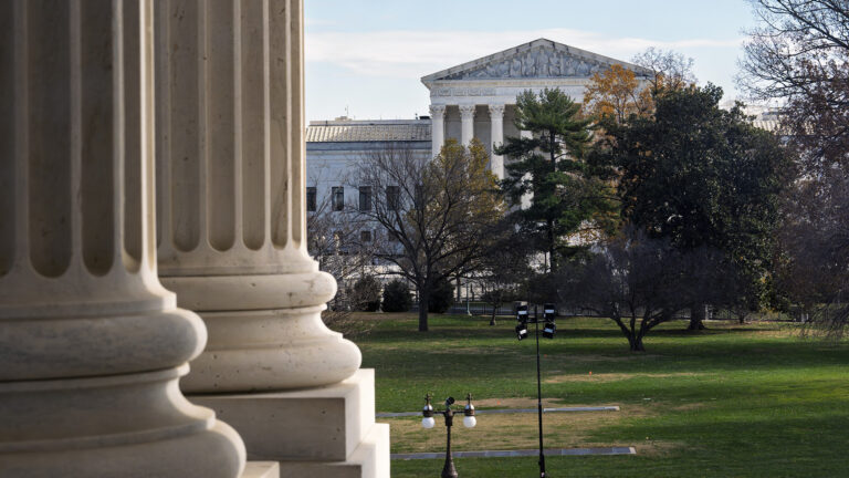 Two marble columns are visible in the foreground in front of a lawn with patches of dead grass and multiple trees with leaves in varying colors in front of a façade of the U.S. Supreme Court Building with marble masonry, a portico of columns, and a pediment with a of sculptures of figures.