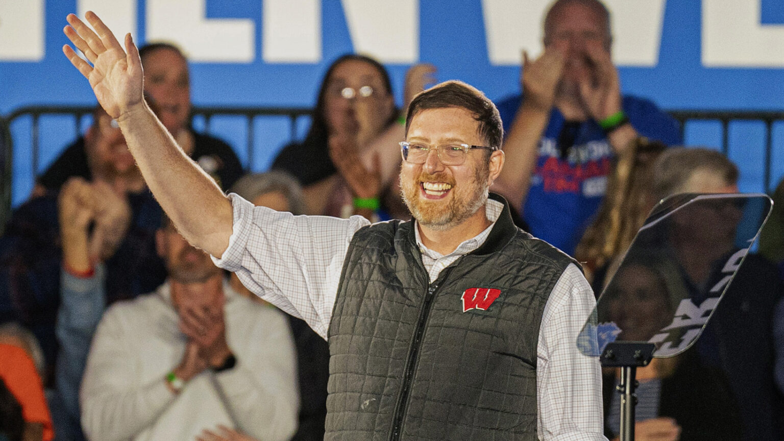 Ben Wikler holds his right hand up and waves while standing behind a teleprompter mirror, with out-of-focus people standing and applauding in the background.