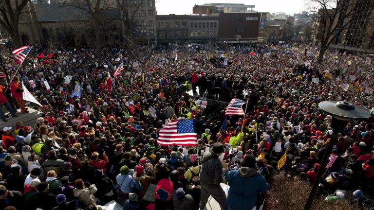 Crowds of people holding signs and flying U.S. flags fill an area bounded by multiple intersecting streets with multi-story buildings.