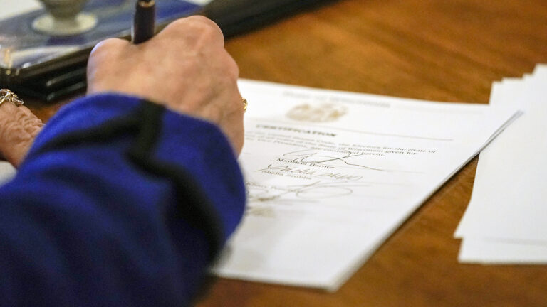 A hand uses a pen to sign a document with multiple other signatures on top of a wood table.