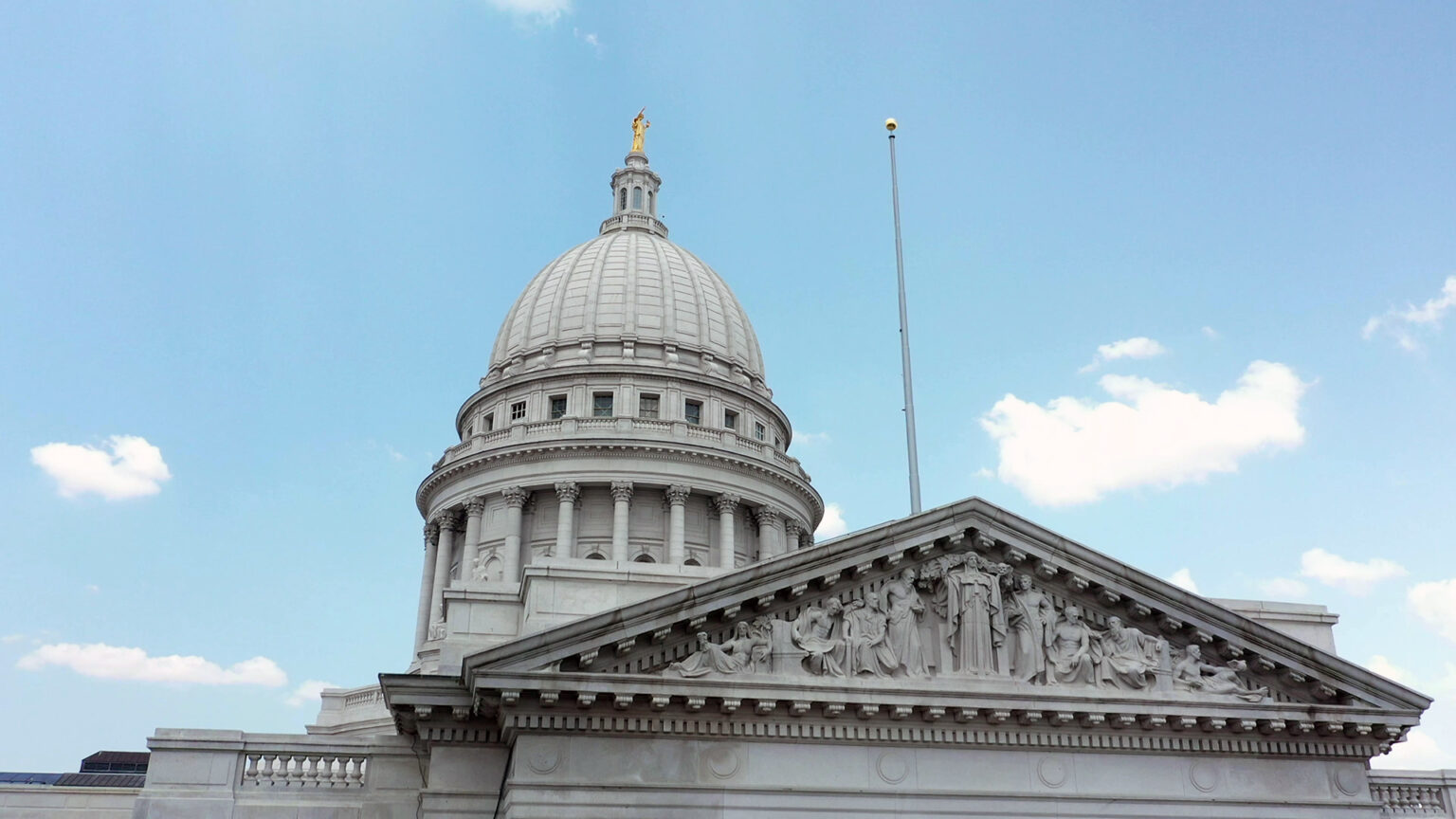 A flagpole is mounted to the top of a pediment on one wing of a marble masonry building with relief statuary and a dome topped by a statue, under a partly cloud sky.