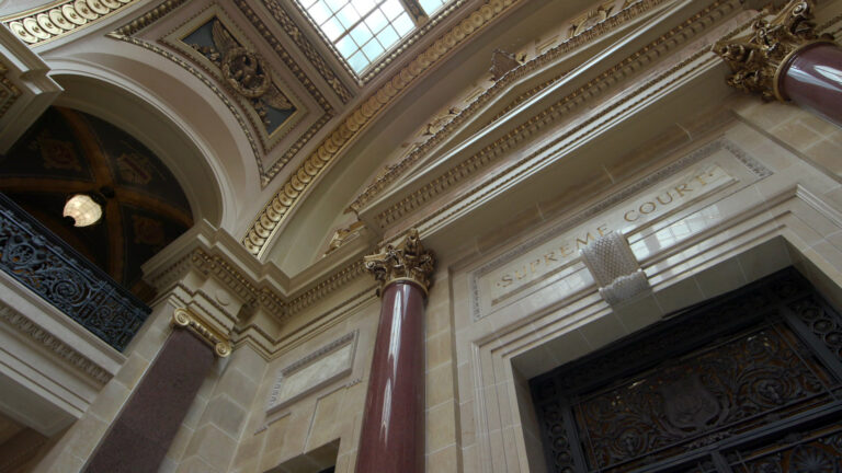 An overhead skylight illuminates marble masonry and two columns with Composite order capitals that flank a doorway with wrought iron filigree, with a carved sign above reading Supreme Court and with a balcony to one side.