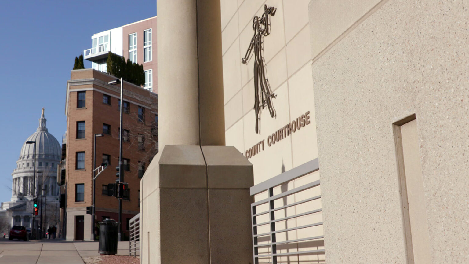 A sign reading County Courthouse and a stylized flat, wire sculpture of Lady Justice are mounted on the masonry wall of a Brutalist-style building, with multi-story brick and metal-sided buildings across an intersection and the Wisconsin State Capitol in the background under a clear sky.