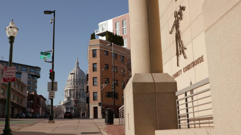 A sign reading County Courthouse and a stylized flat, wire sculpture of Lady Justice are mounted on the masonry wall of a Brutalist-style building, with multi-story brick and metal-sided buildings across an intersection and on either side of a street, with the Wisconsin State Capitol in the background under a clear sky.