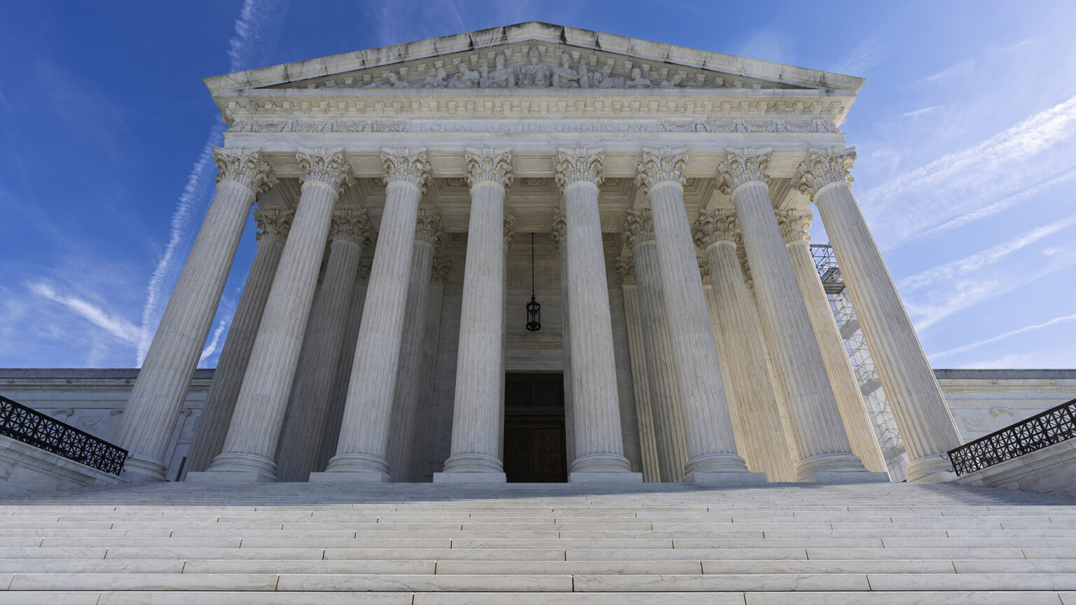 Steps lead to a set of double doors in an entrance on a façade of the U.S. Supreme Court Building with marble masonry, a portico of columns, and a pediment with a of sculptures of figures under a partly cloudy sky.