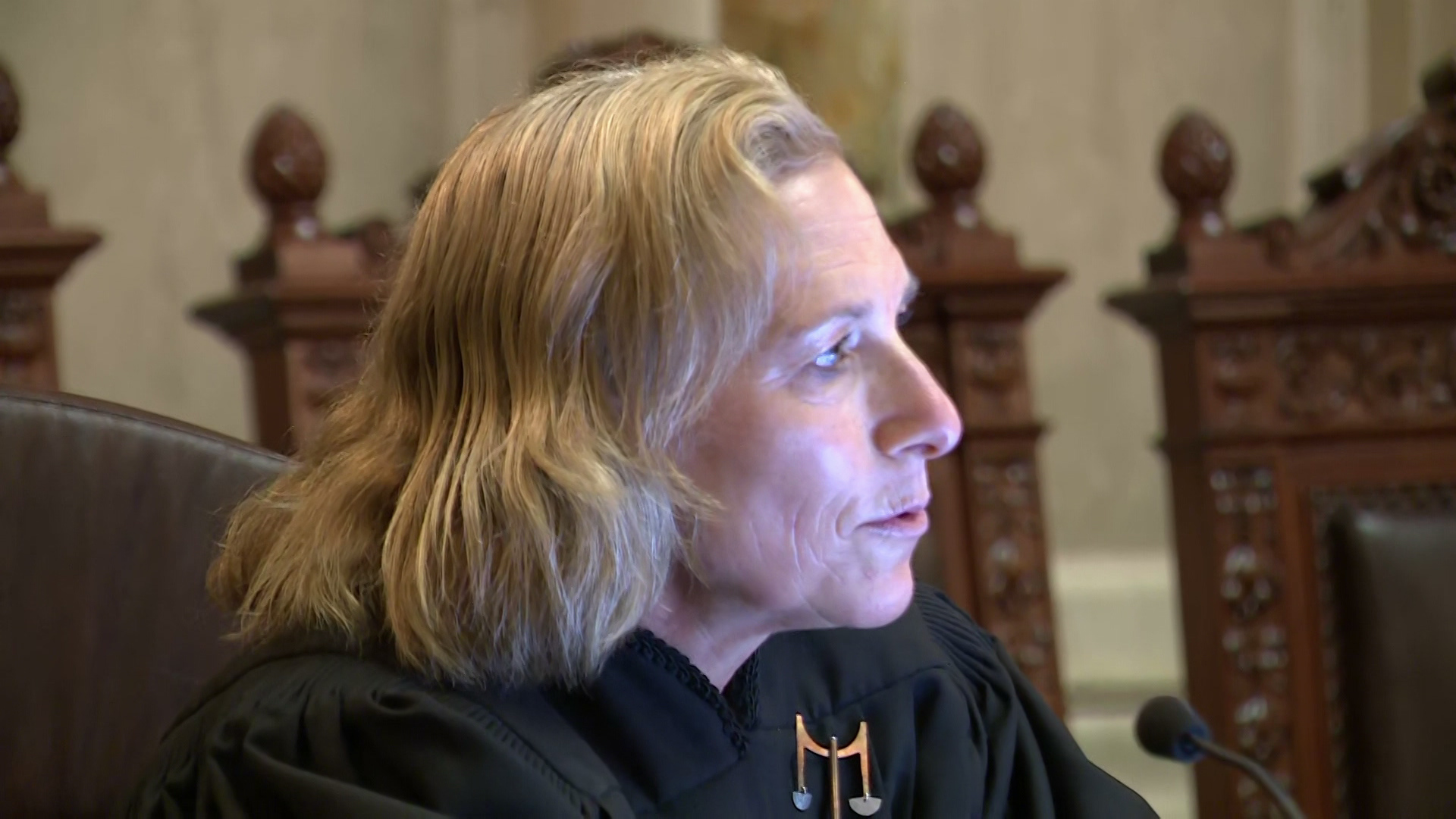 Jill Karofsky speaks into a microphone while seated in a high back leather chairs with other high back leather chairs in the background.