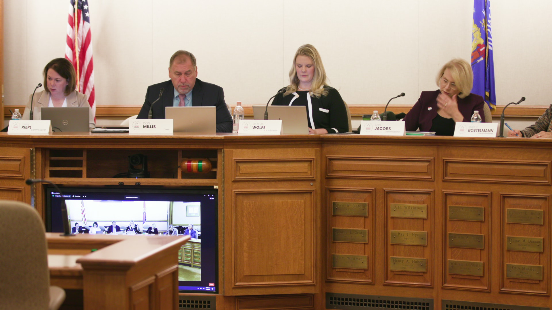 Five people sit behind mounted microphones and paper printed name plates on top of a wood-paneled legislative dais with a TV monitor mounted in the front, with U.S. and Wisconsin flags in the background.