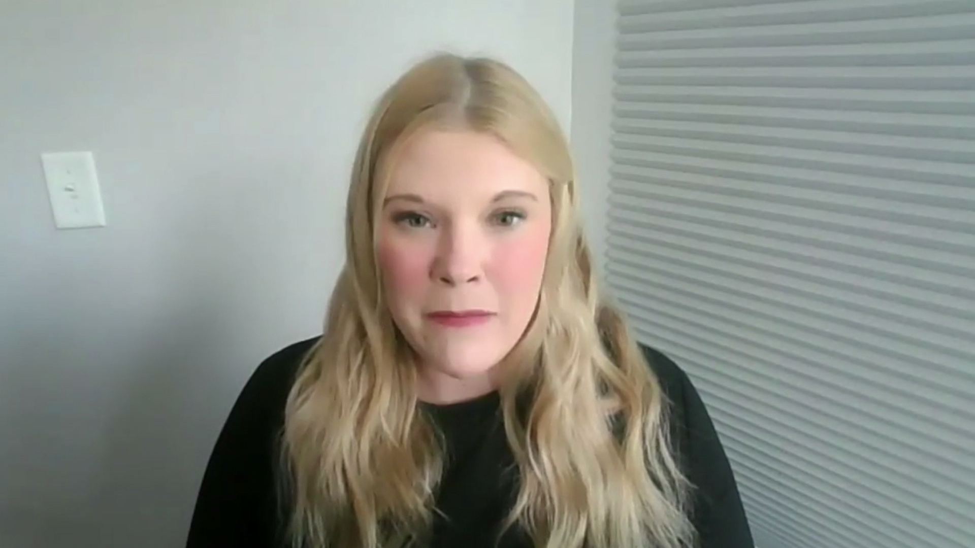 A still image from a video shows Meagan Wolfe speaking while sitting in a room in front of drawn blinds.