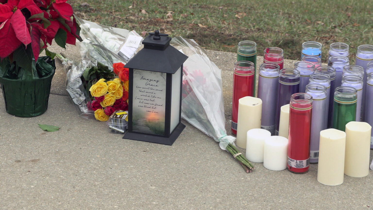 A spontaneous memorial that includes a poinsettia plant, multiple flower bouquets, multiple devotional candles and flameless candles, and an illuminated lanterns with the words Amazing Grace on one of its panels are placed on a concrete sidewalk next to a lawn.