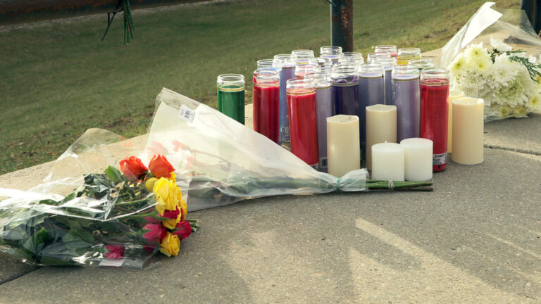 A spontaneous memorial features multiple flower bouquets, multiple devotional candles and flameless candles placed on a concrete sidewalk next to a lawn.