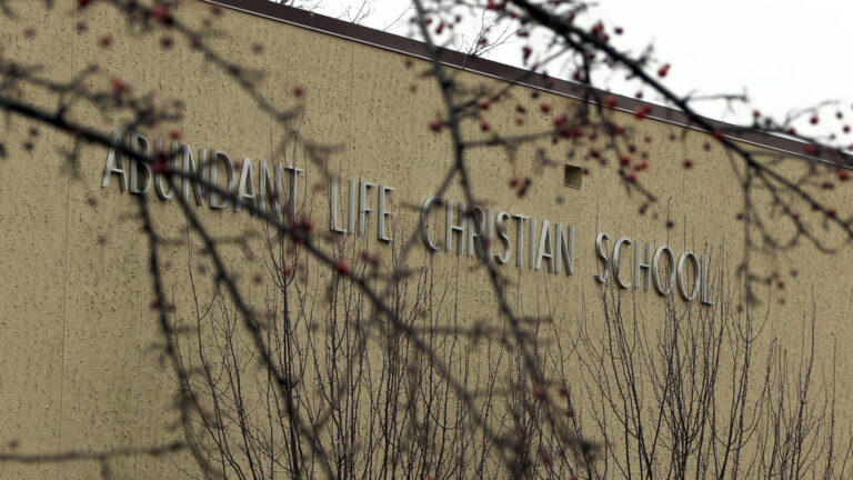 Out-of-focus leafless branches with berries partially obscure a letter sign reading Abundant Life Christian School that is on the side of a stucco wall of a building.