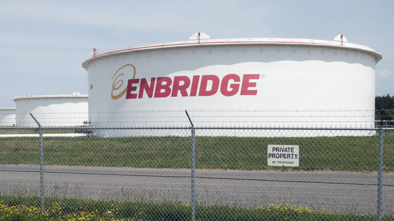 A chain link fence topped by barbed wire and a sign reading Private Property and No Trespassing stands in front of a gravel road and a row of industrial oil storage tanks with the Enbridge wordmark painted on the side of the one in front.