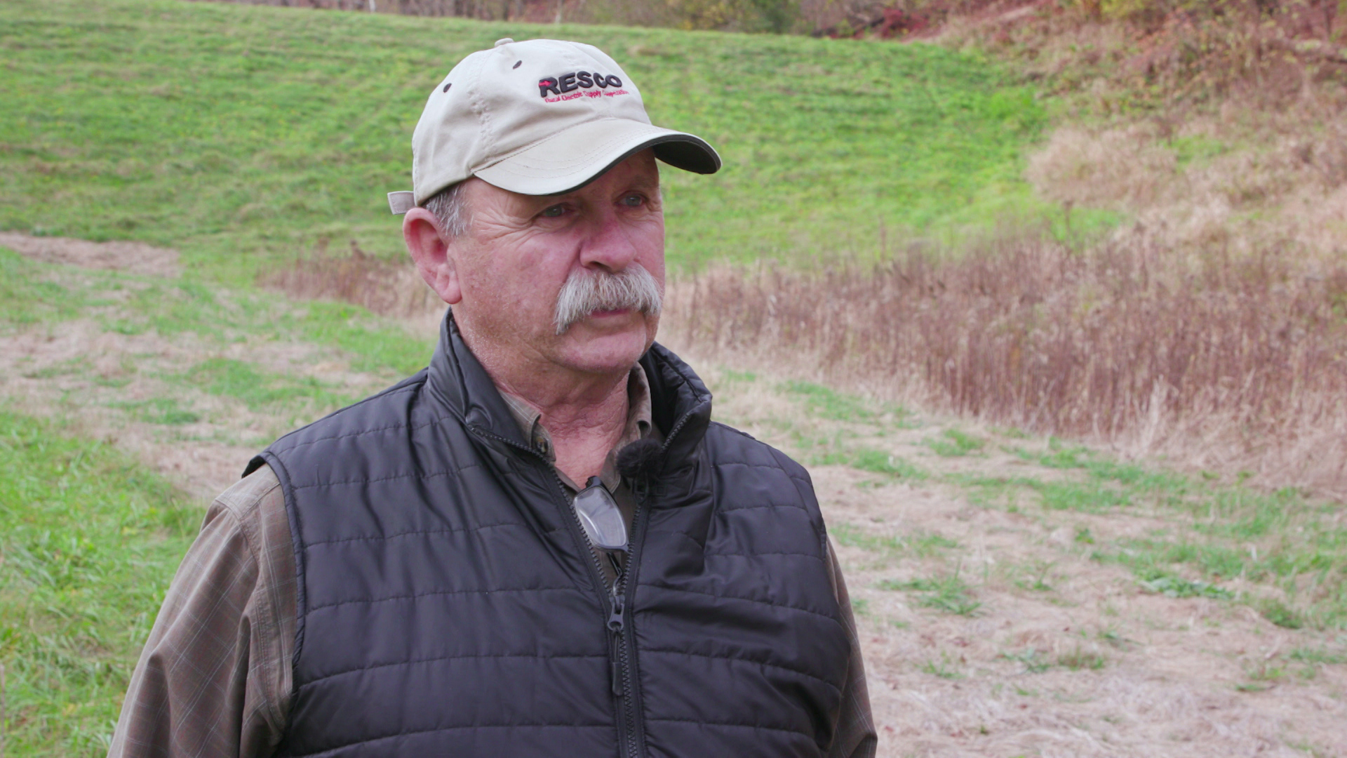 Dave Eggen speaks while standing outdoors with a grass covered earthen dam and other foliage in the background.