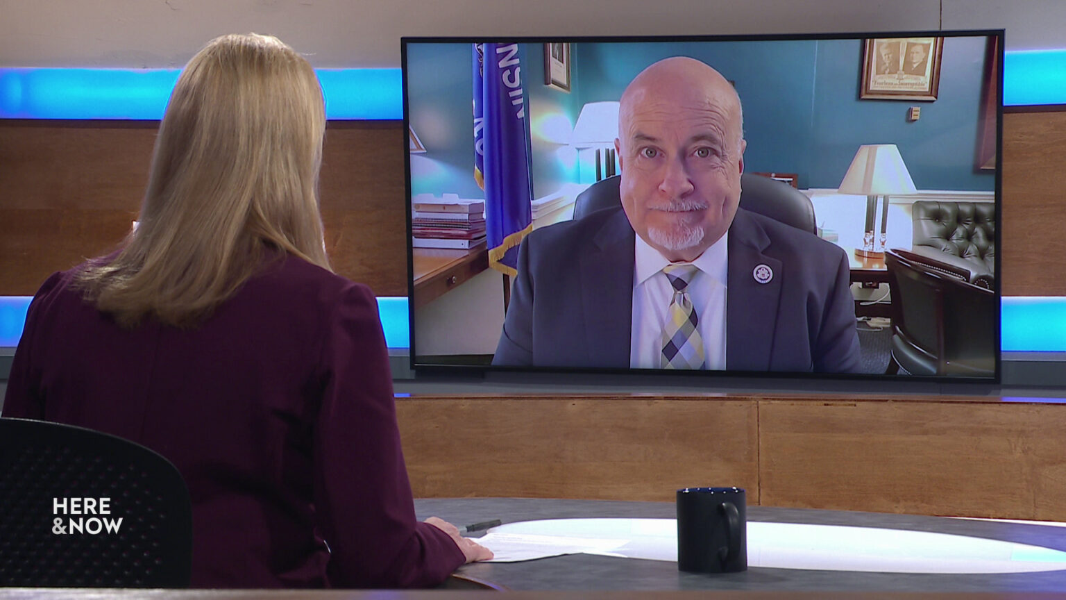 Frederica Freyberg sits at a desk on the Here & Now set and faces a video monitor showing an image of Mark Pocan.