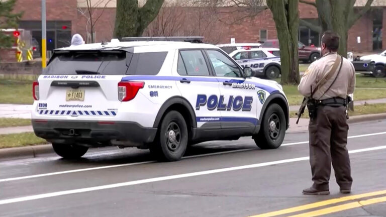 A law enforcement officer wearing a sheriff's deputy uniform and carrying a long gun on a sling around his shoulder stands in the middle of a street next to a parked Madison Police Department vehicle, with other law enforcement vehicles parked on a lawn among tree trunks in front of a brick building with multiple doors and windows.