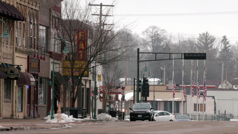 Vehicles are parked on the side and drive along a road with small piles of plowed and shoveled snow, with two-story storefront building on one side next to traffic signals at an intersection, with a row of U.S. flags on flagpoles in font of other buildings, trees and utility lines in the background.