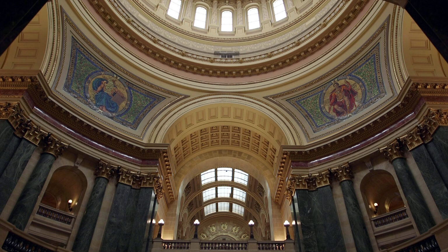 Two mosaics depicting Classical figures — one holding a tablet and labeled Legislation and the other holding staffs and labeled Government — are seen in two quadrants of the interior walls of a rotunda inside a building with marble pillars and other masonry in a Beaux-Arts design.