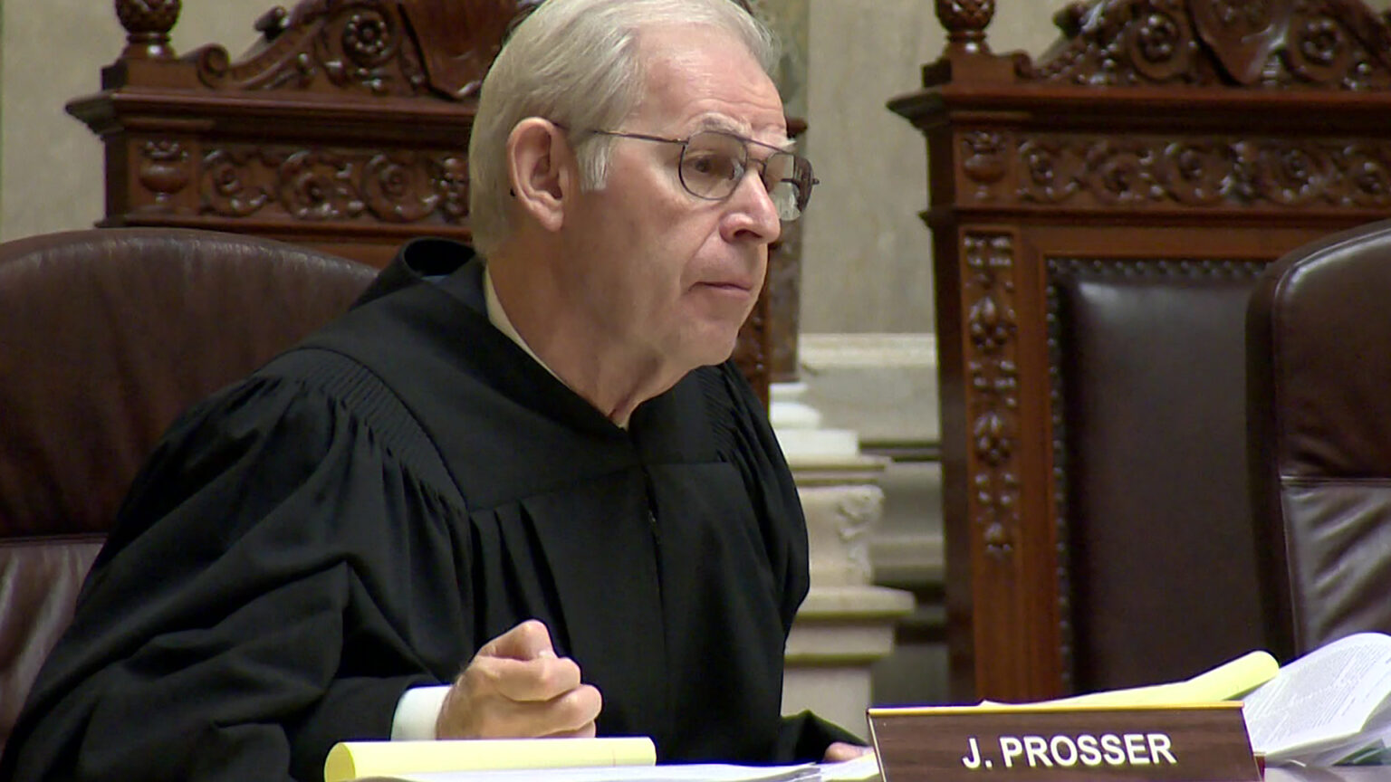 David Prosser sits in a high-backed leather chair on a judicial dais, with multiple papers and a nameplate reading J. Prosser on its surface, with multiple wood and leather high-backed chairs in the background, in a room with marble masonry.