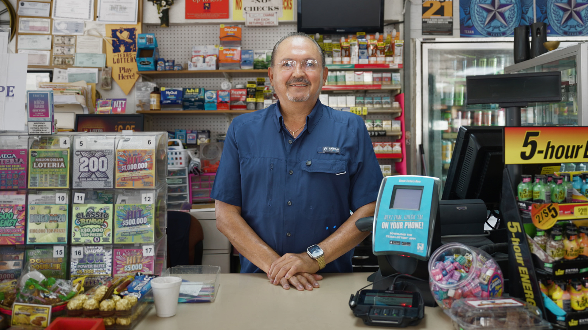 Sergio Garza Castillo stands behind a counter with his hands folded on its surface alongside displays of lottery tickets, energy drinks and candy, as well as a phone payment reader and electronic cash register with monitor, price display and receipt printer, with shelves displaying over-the-counter medications, cigarettes and cigars in the background next to a cooler stocked with beverages.