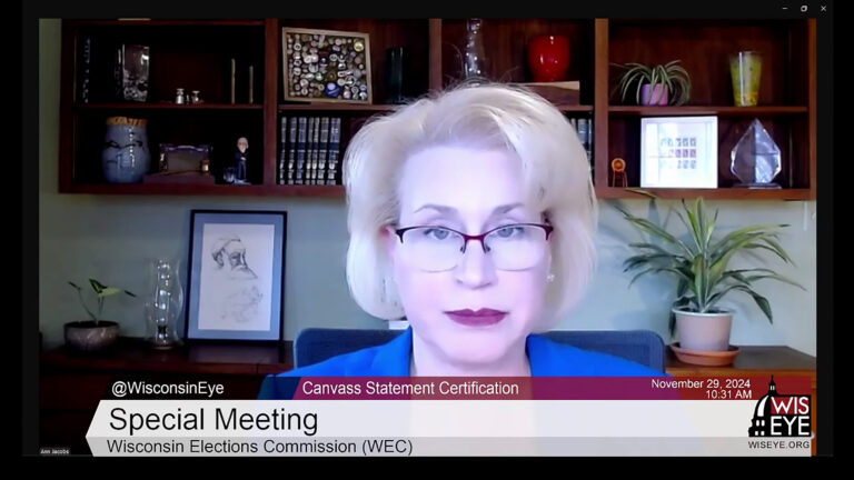 A video still image shows Ann Jacobs sitting in an office with wood shelves filled with books, plants and other items in the background, with a video graphic at bottom including the text Special Meeting and Canvass Statement Certification.