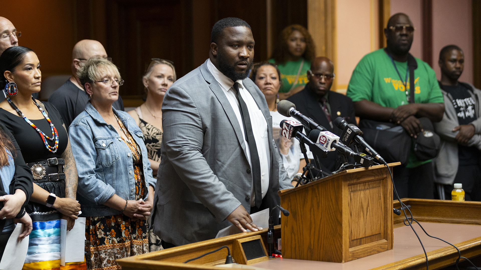 Darrin Madison speaks into multiple microphones with the flags of different media organizations attached to a wood podium, with multiple people standing behind him in the background.