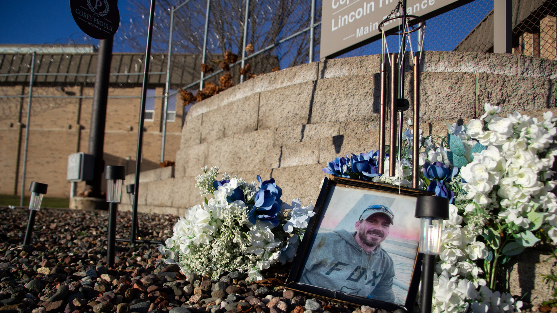 A photo of Corey Proulx and multiple flower bouquets stand among landscaping rocks and in front of a brick terrace, with a two-story chain-link fence topped with two rows of razor wire and multiple buildings in the background.