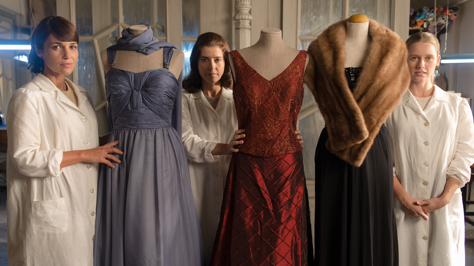 Three women dressed in lab coats stand in a row, each of them next to a dress form covered in elegant evening gowns, including a purple dress, red velvet dress and a black dress draped with a fur stole.