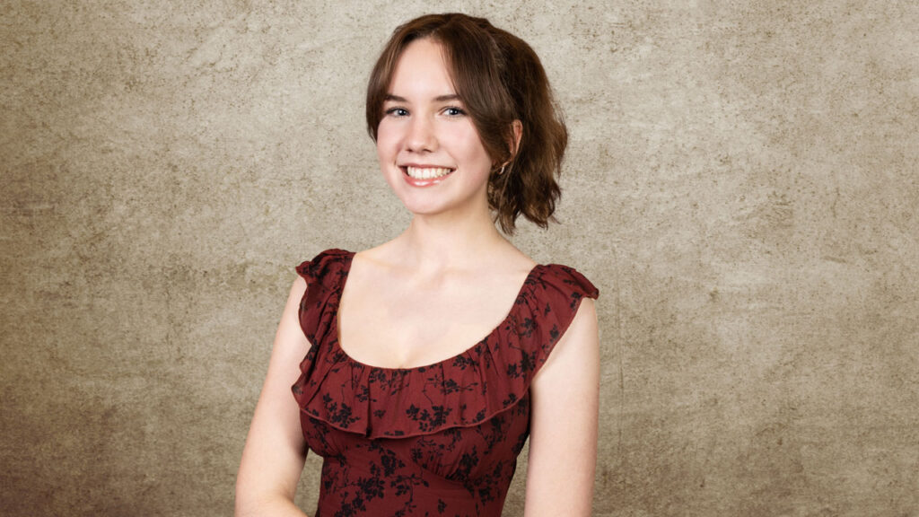 A young teenage girl in a formal dress smiles standing in a portrait studio.