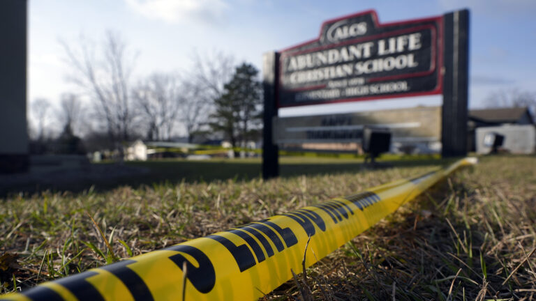 Plastic police tape sits on a lawn in front of an out-of-focus wood sign for Abundant Life Christian School, with out-of-focus trees and buildings in the background.