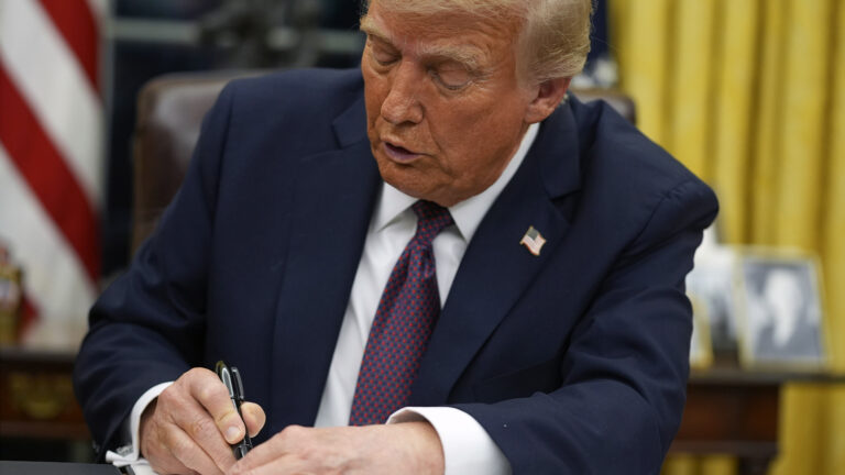 Donald Trump signs a document while sitting in a high-backed leather chair in a room with the stripes of a U.S. flag, curtain and framed photo in the background.