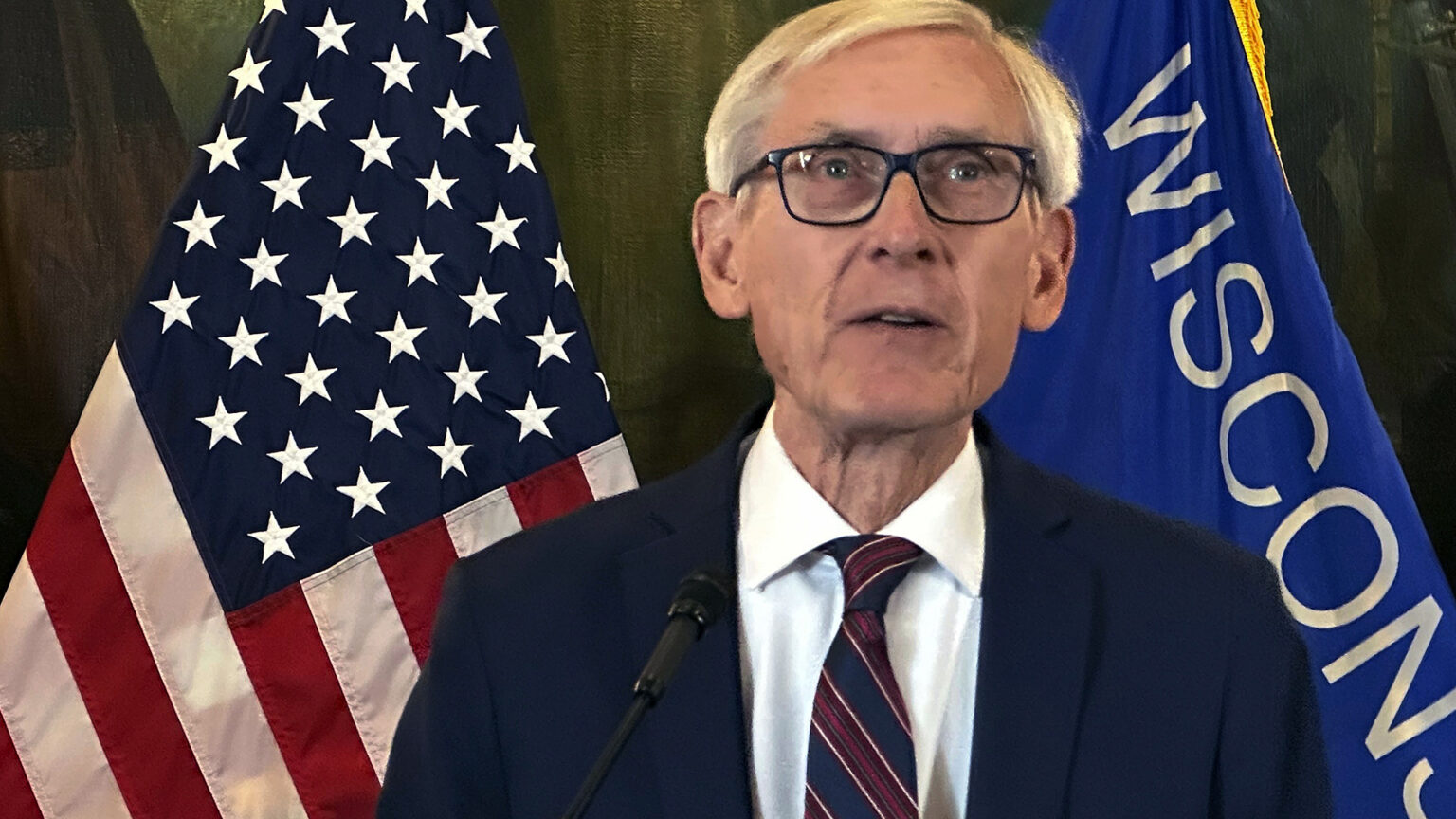 Tony Evers speaks into a mounted microphone while standing in a room with the U.S. and Wisconsin flags in front of a painting in the background.