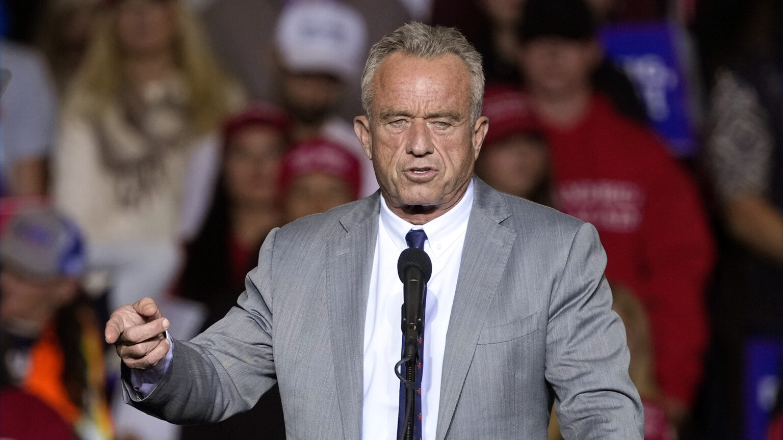 Robert F. Kennedy Jr. points with his right index finger while standing and speaking into a microphone, with out-of-focus people standing in the background.