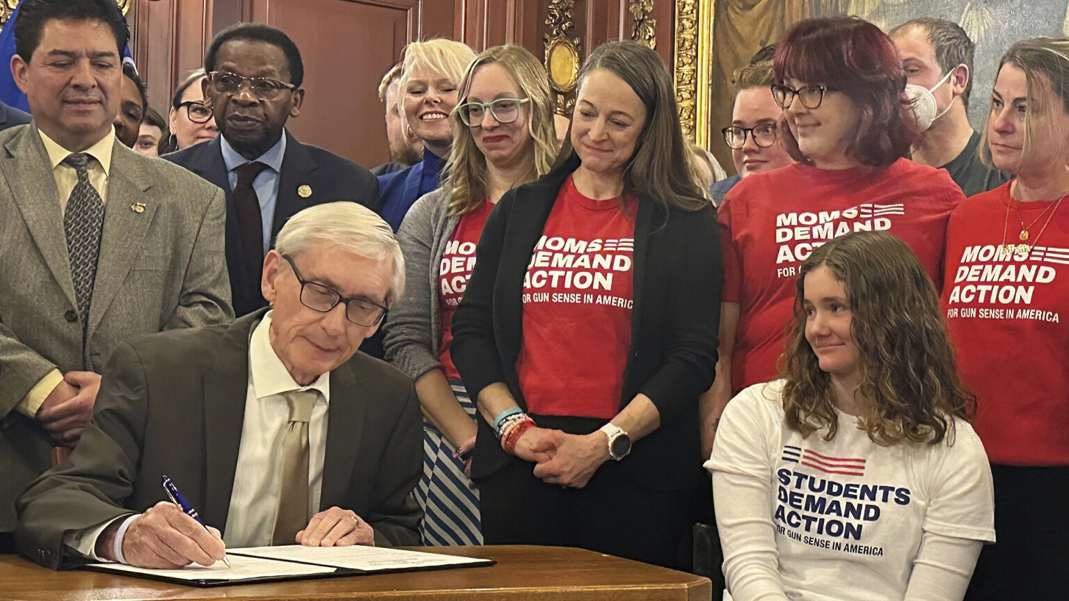 Tony Evers sits at a wood table and uses a pen with his right hand to sign a piece of paper in a folder sitting on its surface, with another person seated next to the table and other people standing behind him in a room with wood-paneled walls and a painting in the background.