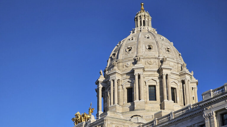 Sunlight reflects off the dome of a Beaux-Arts style building with marble masonry and multiple gold statutes under a clear sky.