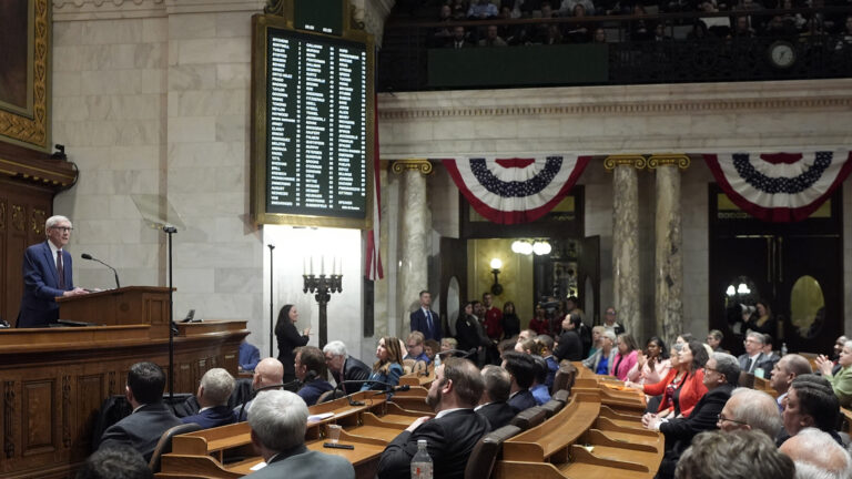 Tony Evers speaks into a microphone mounted to a podium at the center of a wood legislative dais with carved front panels, with people seated at multiple rows of wood desks facing him and other  people standing in the background in front of two sets of double doors with oval windows, in a room with marble masonry and pillars framing red, white and blue bunting, a digital vote register mounted on a wall, illuminated brass light fixtures, and people seated in a gallery in an upper level.