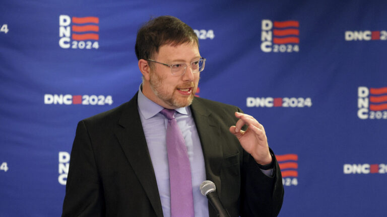 Ben Wikler gestures with his left hand while speaking into a microphone and standing in front of a logo banner with two different graphics with the words DNC and 2024.