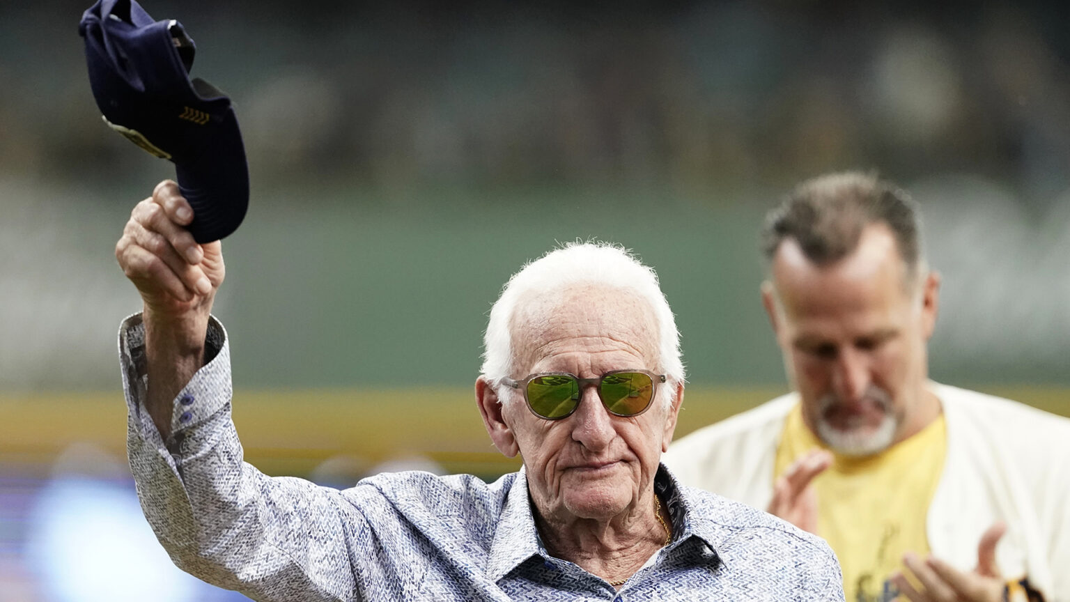 Bob Uecker holds a Milwaukee Brewers baseball cap up in the air with his right hand, with an out-of-focus person standing behind him applauding.