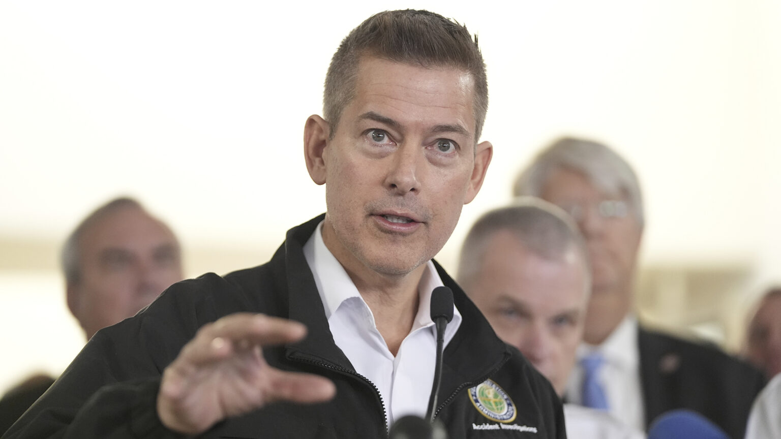 Sean Duffy gestures with his right hand while speaking outdoors, with out-of-focus people standing behind him.