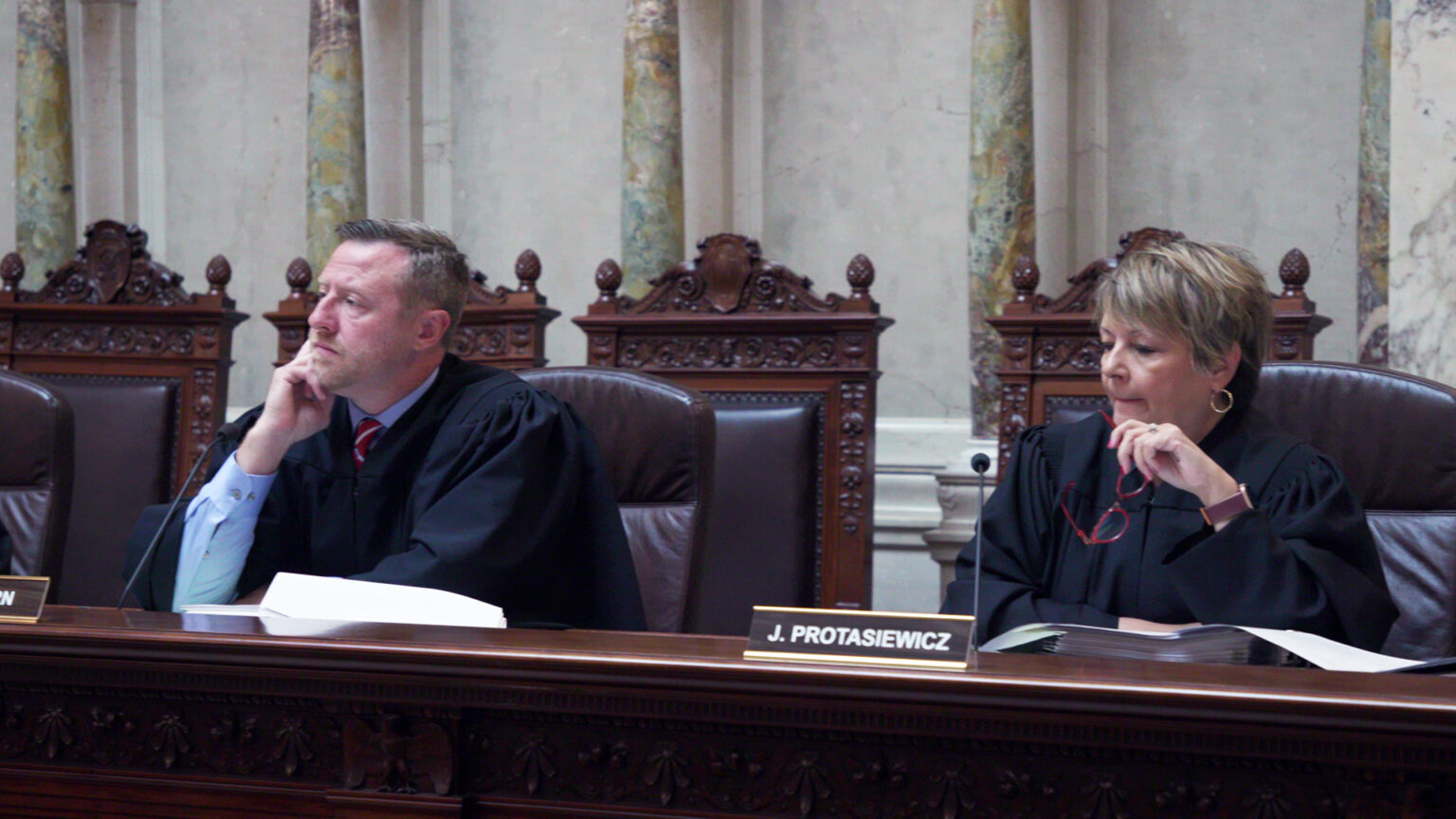 Brian Hagedorn and Janet Protasiewicz sit in high-backed leather chairs at a judicial bench with a nameplate reading J. Protasiewicz, with a row of empty high-backed wood and leather chairs behind them in a room with marble pillars and masonry.