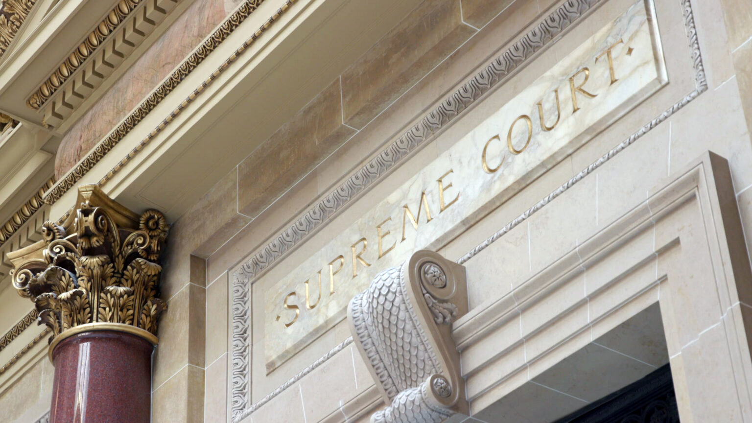 A carved sign reading Supreme Court and a carving of a scroll with scales stands above a doorway, with a marble pillar with a Composite order capital to one side under a marble pediment.